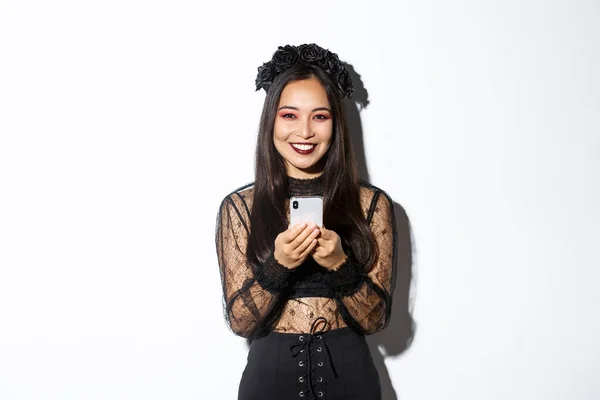 Imagen de la bonita bruja morena elegante en vestido de encaje gótico usando el teléfono móvil, sonriendo a la cámara, de pie sobre fondo blanco — Foto de Stock