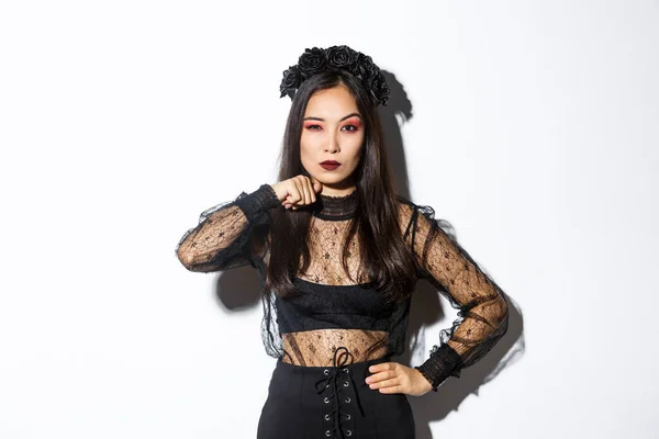 Imagem de bela mulher asiática gótica em vestido de renda preta, ameaçar alguém, fazer gesto de garganta corte e olhando sério para a câmera, celebrando halloween, de pé sobre fundo branco — Fotografia de Stock