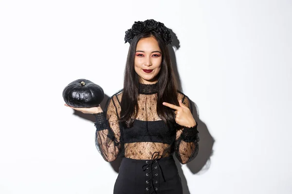 Imagen de una hermosa mujer asiática en vestido de encaje gótico y corona apuntando con el dedo a la calabaza negra, celebrando halloween, vistiendo traje de bruja, de pie sobre fondo blanco —  Fotos de Stock