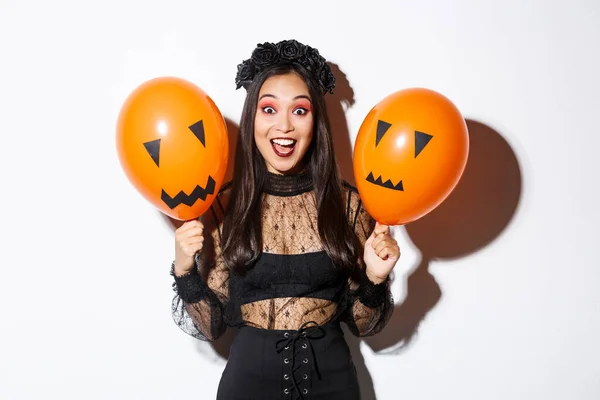 Imagen de mujer asiática feliz en traje de bruja celebrando halloween, sosteniendo globos con caras aterradoras, de pie sobre fondo blanco — Foto de Stock