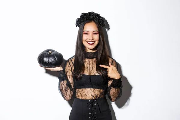 Mutlu gülümseyen Asyalı kadın cadılar bayramında eğleniyor, cadı kostümü giyiyor ve beyaz arka planda duran siyah balkabağını işaret ediyor. — Stok fotoğraf