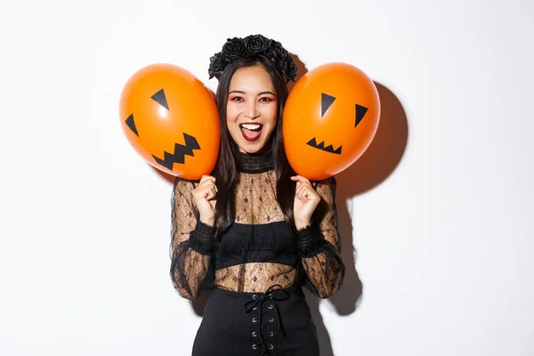 Imagen de chica asiática en traje de bruja malvada sosteniendo dos globos naranjas con caras aterradoras, celebrando halloween, de pie sobre fondo blanco — Foto de Stock