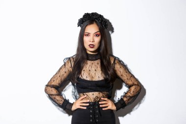 Angry and pissed-off asian female magician, evil witch in black dress and wreath looking mad at someone, frowning and looking judgemental, standing over white background
