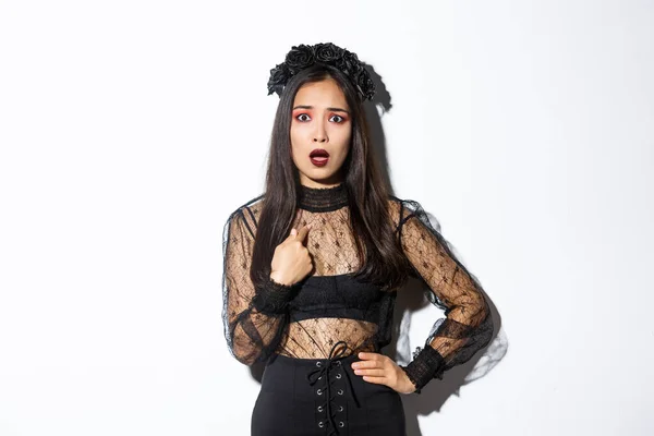 Insultada y confundida mujer asiática en traje de halloween apuntándose a sí misma, vistiendo vestido negro de bruja malvada en la fiesta, de pie sobre fondo blanco —  Fotos de Stock