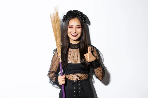 Imagen de mujer asiática sonriente en traje de bruja con escoba, mostrando pulgares hacia arriba en aprobación, de pie sobre fondo blanco —  Fotos de Stock