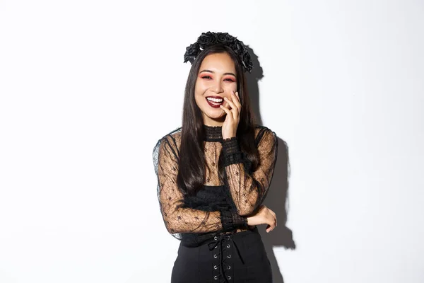 Изображение счастливой красивой азиатки в готическом платье и черном венке, смеющейся на вечеринке в честь Хэллоуина, стоящей на белом фоне — стоковое фото