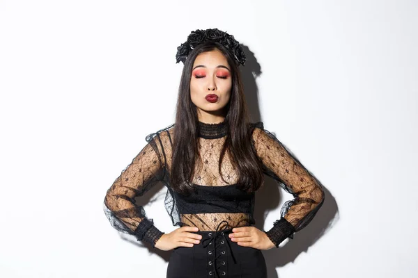 Imagen de la hermosa chica asiática en traje de halloween cerrar los ojos y pucheros, esperando un beso, de pie sobre fondo blanco con vestido negro gótico con corona de rosas —  Fotos de Stock