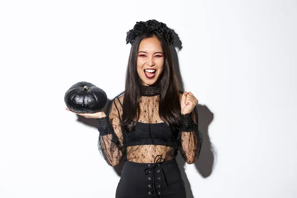 Aantrekkelijk vrolijk aziatisch meisje genieten van halloween, het houden van zwarte pompoen en juichen, het dragen van heks kostuum, staande over witte achtergrond — Stockfoto