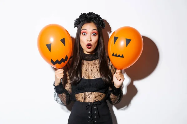 Imagen de mujer asiática sorprendida en traje de bruja celebrando halloween, sosteniendo globos con caras aterradoras, de pie sobre fondo blanco — Foto de Stock