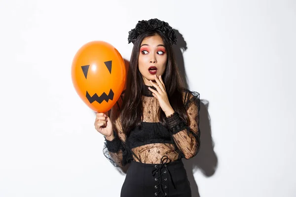 Portrait of girl looking scared at orange balloon with creepy face, wearing witch costume, celebrating halloween — Stock Photo, Image