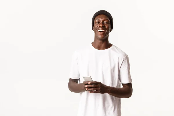 Portrait d'attrayant homme afro-américain insouciant en bonnet riant et souriant, en utilisant l'application de téléphone mobile, fond blanc — Photo