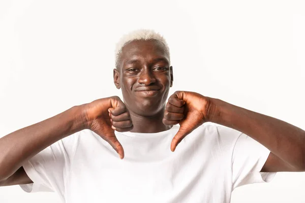 Close-up of unamused and skeptical Black guy with blond hair, showing thumbs-down and smirk with dislike, disagreed bad thing, standing white background — Stok Foto