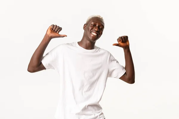 Retrato de un atractivo estudiante rubio afroamericano confiado apuntándose a sí mismo y sonriendo orgulloso, de pie sobre un fondo blanco —  Fotos de Stock