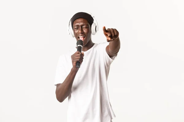 Retrato de un joven afroamericano despreocupado y fresco, actuando en karaoke, cantando en el micrófono mientras usa auriculares, fondo blanco — Foto de Stock