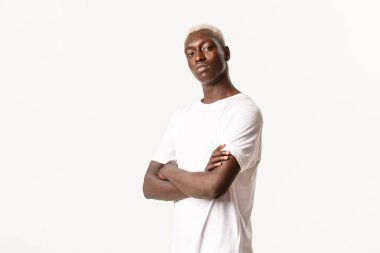 Kendine güveni tam Afro-Amerikan bir adamın portresi. Sarı saçlı, çapraz kollu göğsü olan ve arsız görünen, beyaz arka planda duran.