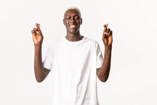 Retrato de esperançoso sorridente afro-americano loiro cara, sorrindo otimista e fazendo desejo, dedos cruzados boa sorte, fundo branco — Fotografia de Stock