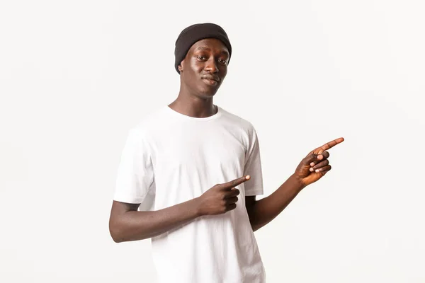 Portret van een lachende aantrekkelijke Afrikaans-Amerikaanse man in muts, wijzende vingers naar rechts, met logo of reclamebanner, staande witte achtergrond — Stockfoto
