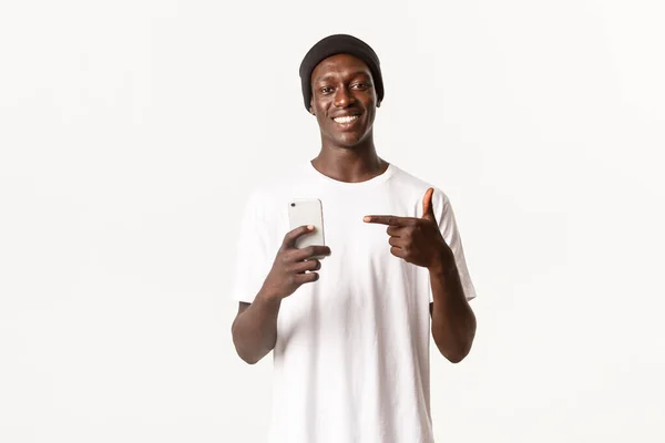 Retrato de un joven afroamericano sonriente que recomienda la aplicación para teléfonos inteligentes, señalando con el dedo el teléfono móvil con la cara satisfecha — Foto de Stock