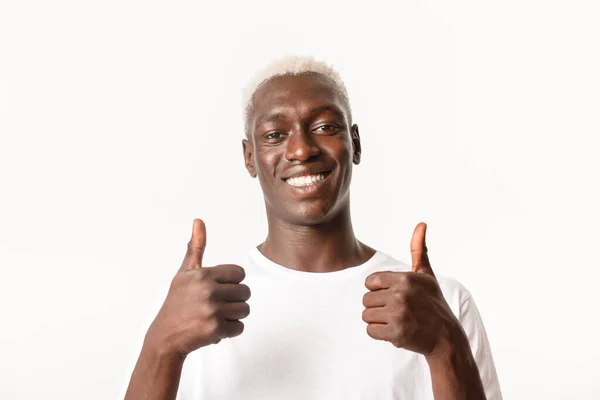 Primer plano de chico rubio afroamericano guapo complacido sonriendo, mostrando pulgares hacia arriba en aprobación, como algo, de pie fondo blanco — Foto de Stock