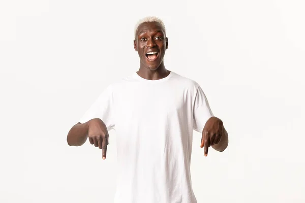 Şaşırmış Afro-Amerikan sarışın adamın portresi, harika bir logo gösteriyor, gülümsüyor ve parmaklarını aşağı işaret ediyor. — Stok fotoğraf