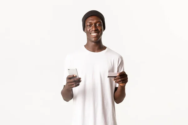 Retrato de chico afroamericano feliz y satisfecho, guapo usando tarjeta de crédito y teléfono móvil para ir de compras en línea o entrar en la cuenta bancaria electrónica, fondo blanco — Foto de Stock
