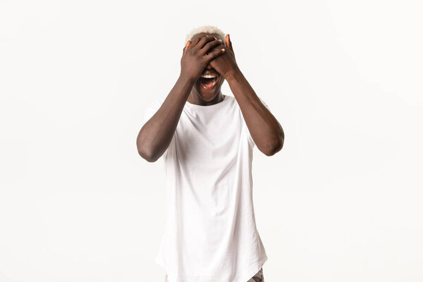 Portrait of surprised and amused african-american blond guy, shut eyes with hands and smiling amazed, white background.