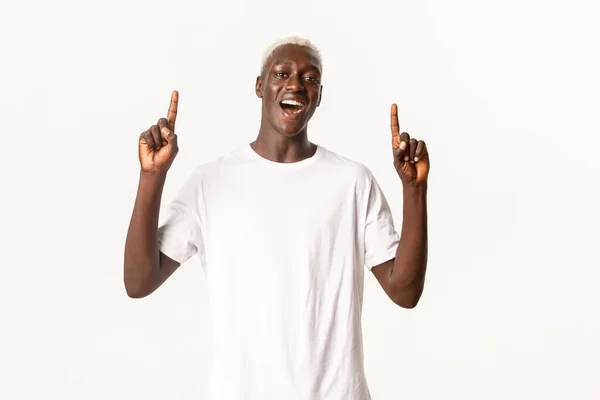 Portret van vrolijke Afrikaans-Amerikaanse blonde man, wijzende vingers omhoog en tonen logo, glimlachend verbaasd, staande witte achtergrond — Stockfoto