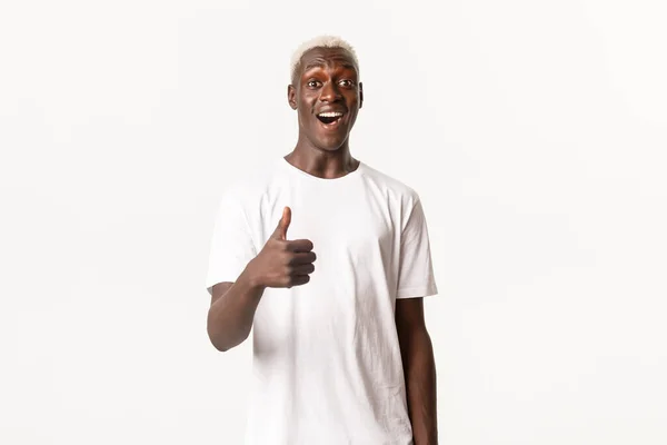Retrato de homem loiro afro-americano sorridente impressionado, mostrando polegares para cima, elogiando boa escolha, fazendo elogios, fundo branco — Fotografia de Stock
