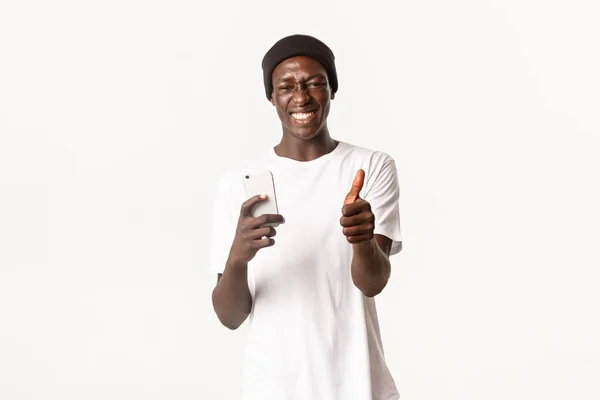 Retrato de chico afroamericano feliz y satisfecho, guapo sonriendo ampliamente, mostrando pulgares hacia arriba después de usar la aplicación de teléfono móvil, fondo blanco — Foto de Stock