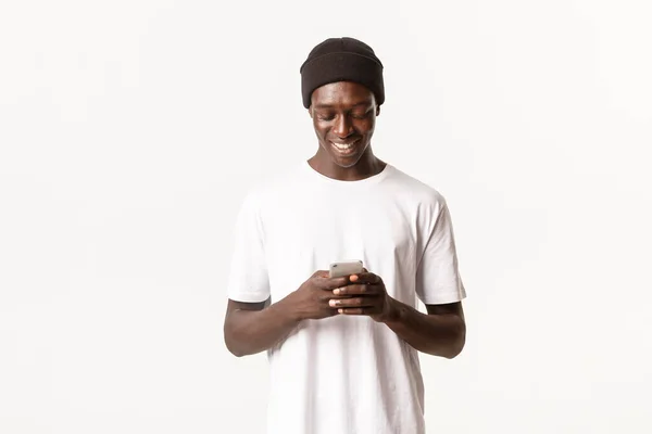 Retrato del joven afroamericano guapo en gorro usando teléfono móvil, mensajería y sonrisa en la pantalla del teléfono inteligente, fondo blanco — Foto de Stock