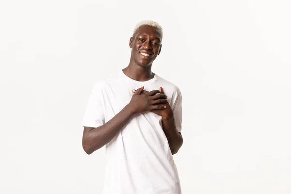 Porträtt av glad och rörd afrikansk amerikansk blond man, leende och hålla händerna på hjärtat glad, stående vit bakgrund — Stockfoto