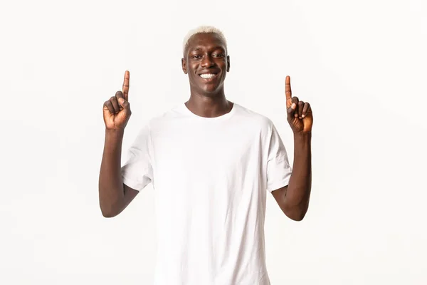 Retrato del alegre hombre rubio afroamericano, señalando con los dedos hacia arriba y sonriendo feliz, mostrando logotipo o anuncio, fondo blanco — Foto de Stock