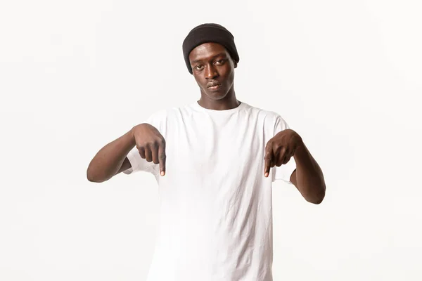 Portrait of serious african-american guy in beanie, looking at camera and pointing fingers down with poker face, white background — Stock Photo, Image