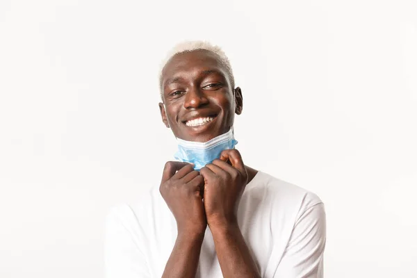 Close-up of handsome, happy african-american blond guy take-off medical mask and smile at camera carefree, standing over white background — Stok Foto
