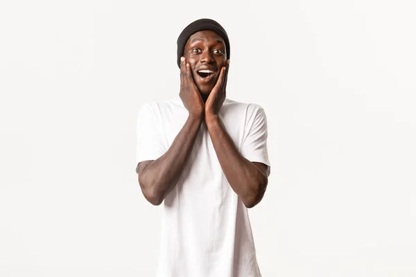 Retrato de feliz sorprendido joven afroamericano chico, mirando asombrado y alegre, jadeando fascinado, cogido de la mano en las mejillas, fondo blanco — Foto de Stock