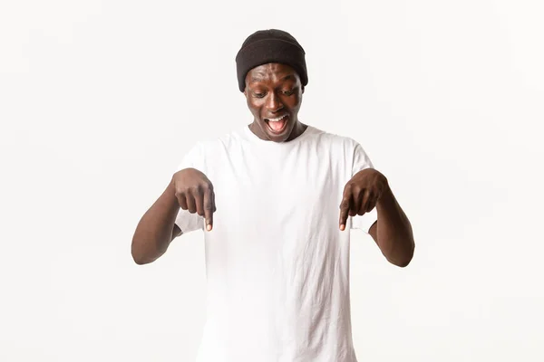 Retrato de hombre afro-americano emocionado y feliz en gorro, gritando de asombro y señalando con los dedos hacia abajo, mirando su logotipo alegre, de pie fondo blanco — Foto de Stock