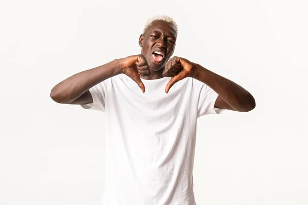 Retrato de un hombre rubio afroamericano disgustado, mostrando la lengua y los pulgares hacia abajo, disgusta y desaprueba algo, de pie fondo blanco — Foto de Stock