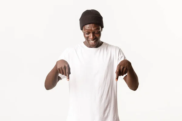 Πορτρέτο του χαρούμενου και διασκεδαστικού Αφροαμερικανού όμορφου άντρα με το σκούφο, να κοιτάζει και να δείχνει με τα δάχτυλα κάτι ενδιαφέρον, λευκό φόντο — Φωτογραφία Αρχείου