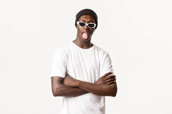 Retrato de tipo afroamericano divertido y fresco en gorro y gafas de sol, mostrando la lengua con expresión sin molestias, de pie fondo blanco — Foto de Stock