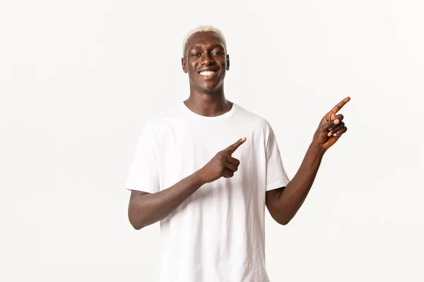 Πορτρέτο του ικανοποιημένου ευτυχισμένος αφρικανός-Αμερικανός ξανθός άνδρας, χαμογελώντας ευχαριστημένος και δείχνοντας τα δάχτυλα πάνω δεξιά γωνία, δείχνοντας το λογότυπο, λευκό φόντο — Φωτογραφία Αρχείου
