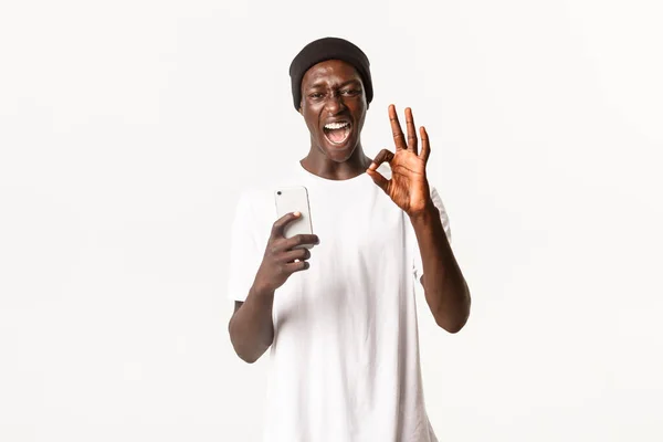 Portré hűvös afro-amerikai fiatal srác mutat rendben gesztus, mint az alkalmazás, a mobiltelefon és elégedett, álló fehér háttér — Stock Fotó