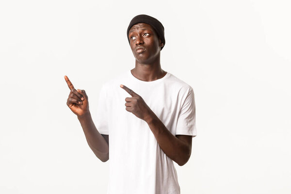 Portrait of upset gloomy african-american guy in beanie, pouting sad, pointing fingers upper left corner something disappointing, white background.