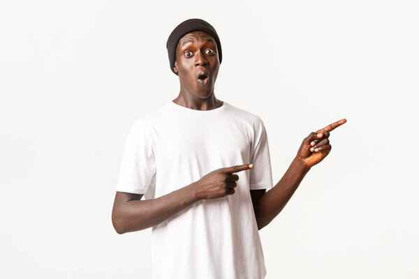 Portrait of impressed and excited african-american man in beanie, pointing fingers right and say wow astonished, standing white background.