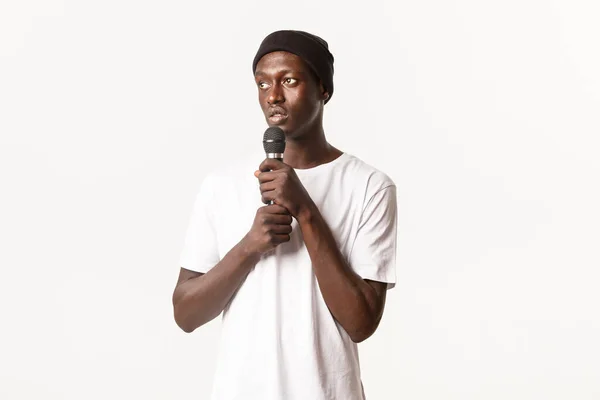 Retrato de un joven afroamericano en gorro, cantando karaoke, mirando a la izquierda y sosteniendo el micrófono, de pie fondo blanco — Foto de Stock