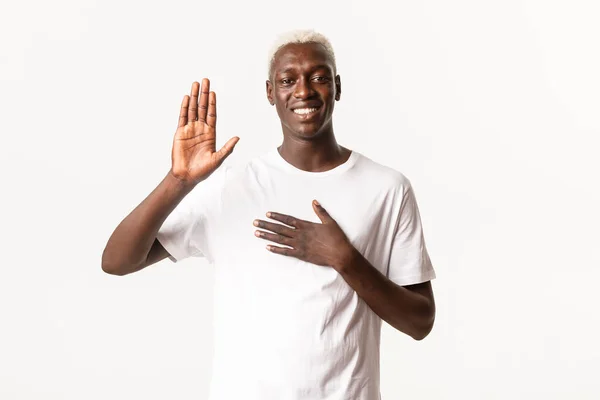 Porträtt av ärlig attraktiv afrikansk-amerikansk blond kille, höja en hand och en annan på hjärtat, ge löfte, ler över vit bakgrund — Stockfoto