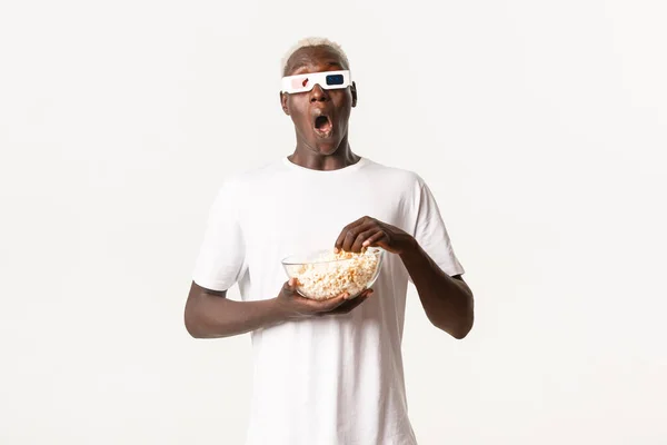 Retrato de chico rubio afroamericano guapo emocionado, viendo una película increíble en gafas 3D, comiendo palomitas de maíz, mirando asombrado sobre fondo blanco — Foto de Stock