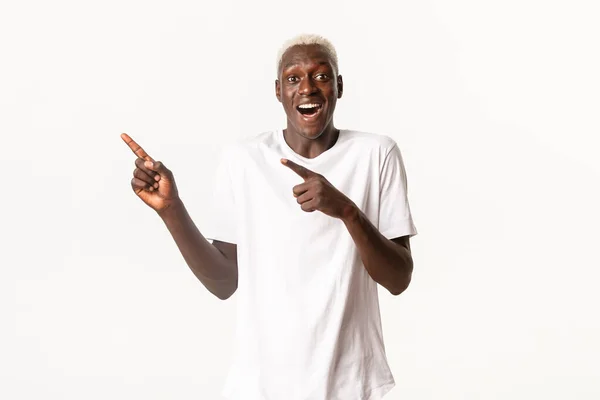 Retrato de emocionado hombre rubio afroamericano feliz, mirando fascinado y señalando los dedos esquina superior izquierda, fondo blanco —  Fotos de Stock