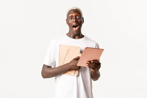 Retrato de estudiante varón rubio afroamericano, guapo y sorprendido, con gafas, portátiles para estudiar y tableta digital, fondo blanco —  Fotos de Stock