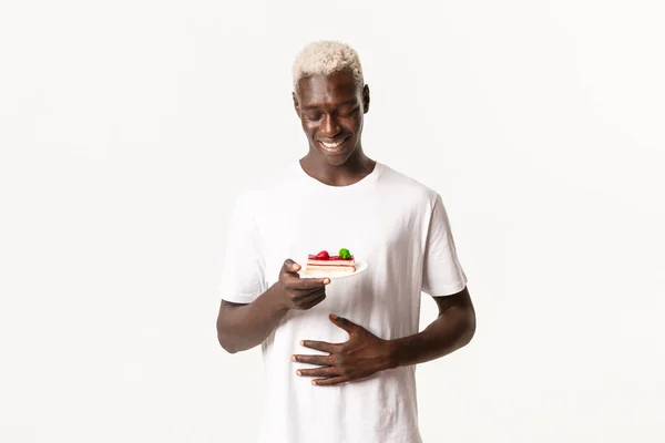 Retrato del joven rubio afroamericano contento y feliz, tocando el vientre satisfecho y mirando deliciosa torta, de pie fondo blanco — Foto de Stock