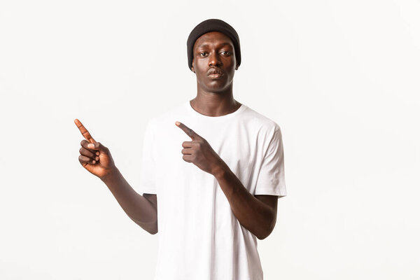 Portrait of serious african-american guy in beanie, pointing fingers upper left corner, showing logo, white background.